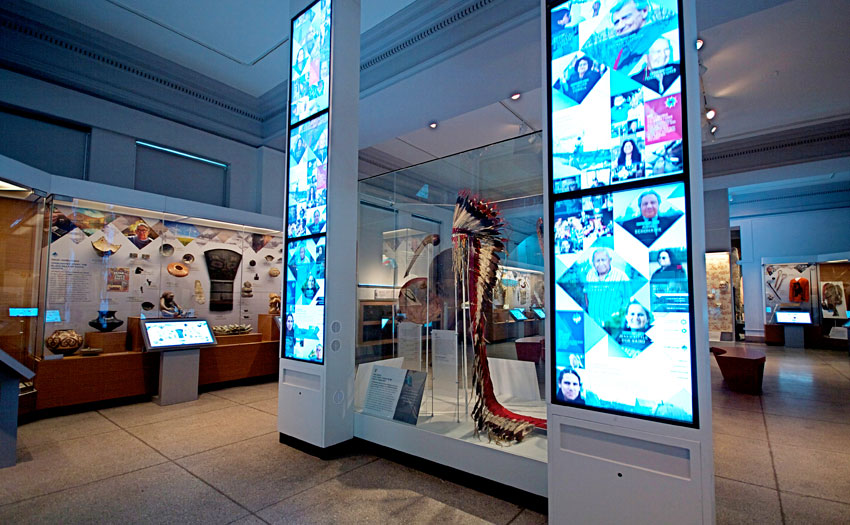 A tall central display case holding a feathered headdress, flanked by vertical screens showing a grid of contemporary Native American faces and voices.