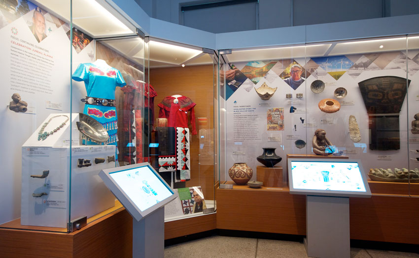 Two display cases holding brightly colored Native American beaded garments and pottery, with touchscreen interactives integrated into the case-front counter.