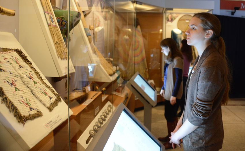 A visitor leaning in to examine a piece of beadwork in a display case, with another visitor at a touchscreen behind her.