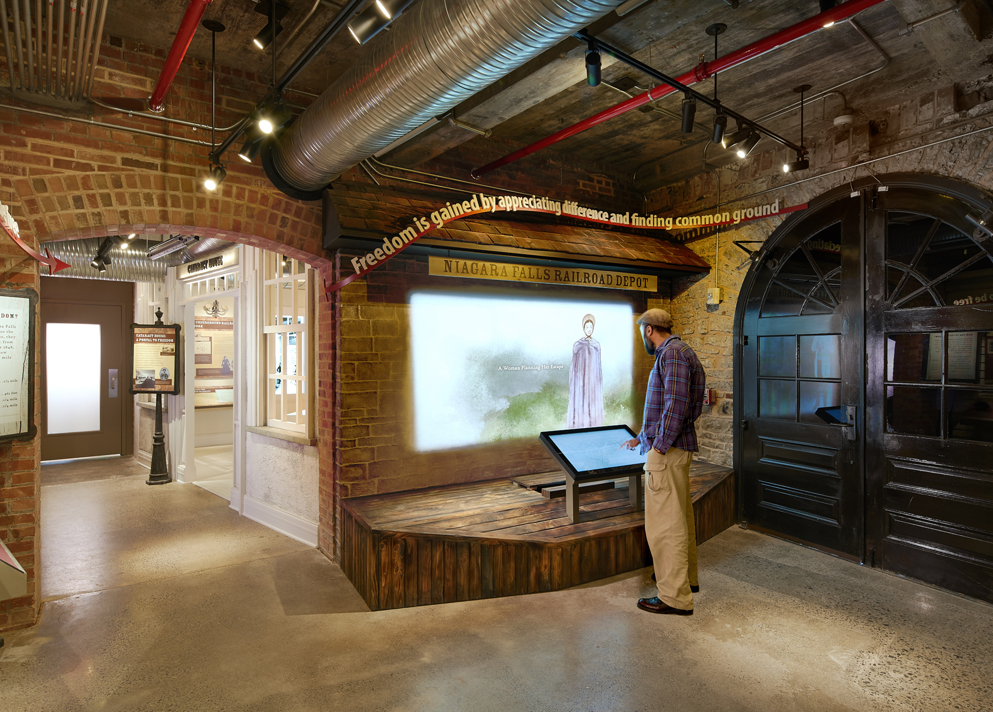 A visitor at a touchscreen kiosk in front of a projected video on a brick wall, with the headline 'Freedom is gained by appreciating difference and finding common ground' above the screen.