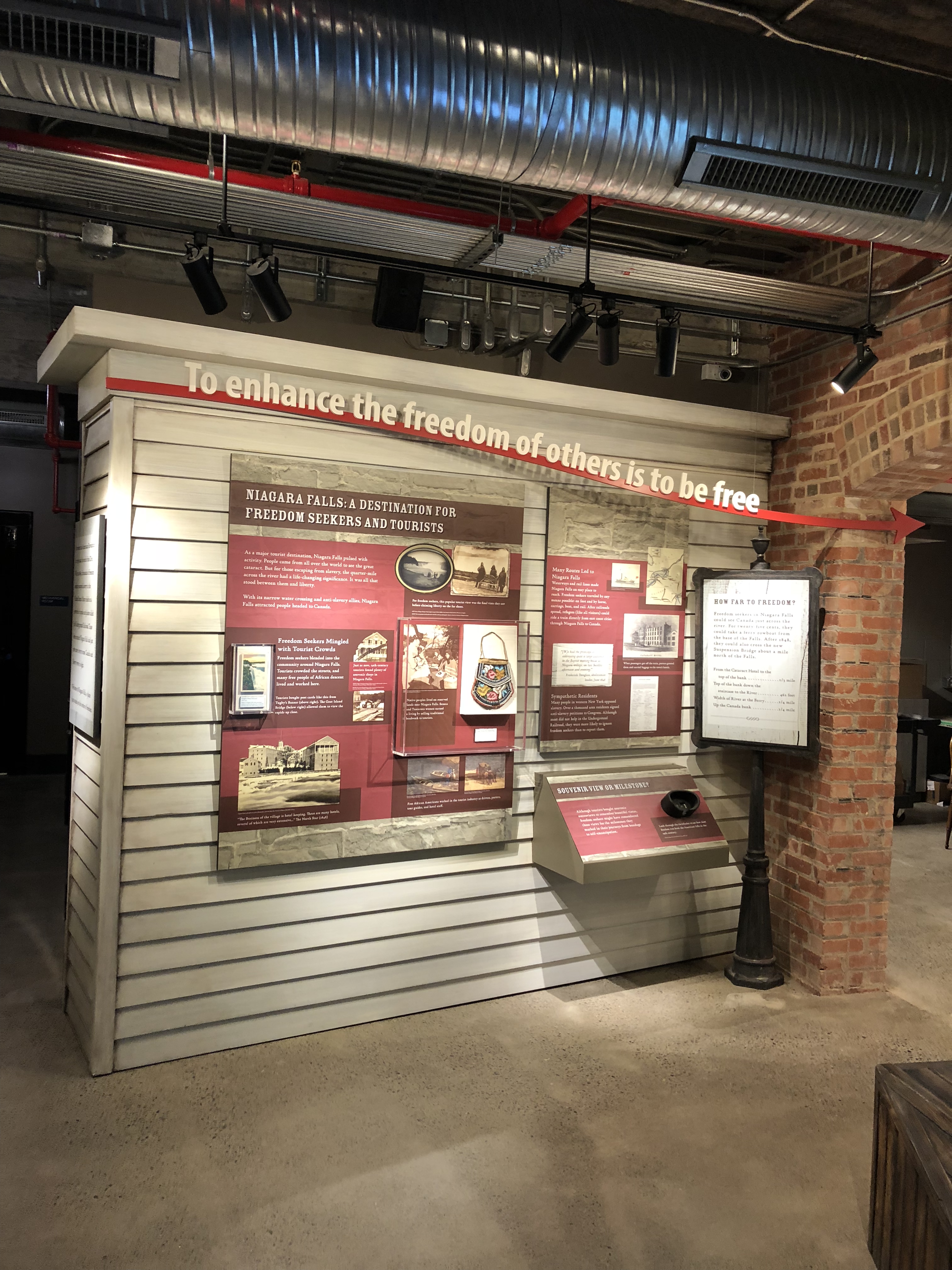 A clapboard depot-style exhibit feature with the message 'To enhance the freedom of others is to be free,' showing interpretive panels and a wayside reader.
